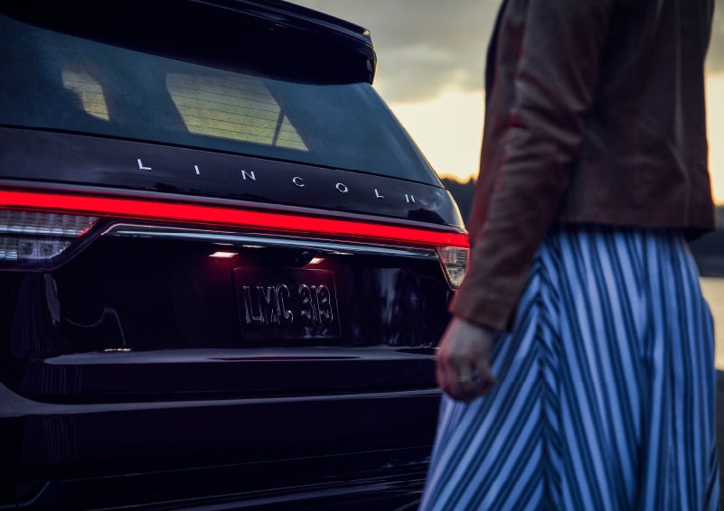 A person is shown near the rear of a 2024 Lincoln Aviator® SUV as the Lincoln Embrace illuminates the rear lights | Sheehy Lincoln of Richmond in Richmond VA